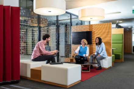 Three MeteoSwiss employees, two women and a man, sit on white sofa stools in the MeteoSwiss offices and discuss with each other over a coffee. A laptop is placed on the table between them.